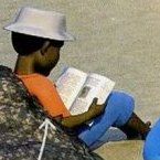 A boy in a white hat sitting on a rock and reading a book