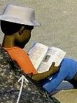 A boy in a white hat reading a book on the beach