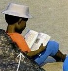 A boy in a white hat sitting on a rock and reading a book