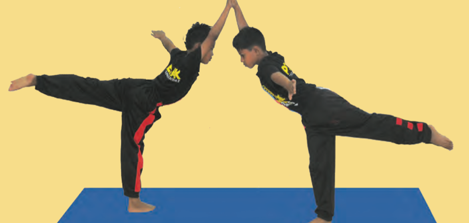 Dua murid berdiri pada satu kaki, kaki sebelah lagi lurus ke belakang, tangan bertemu di atas