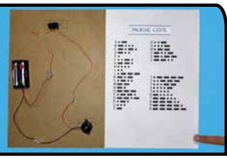 A finished telegraph machine: dry cell holder, wires, buzzer and switch on cardboard beside a Morse code chart