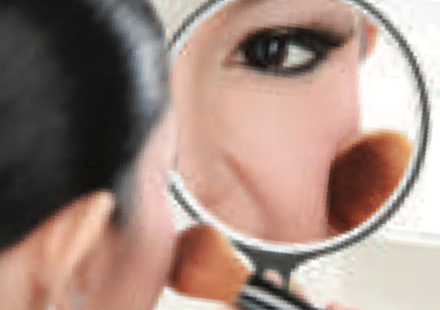 A woman applying powder while looking into a hand mirror that shows an enlarged image of her eye