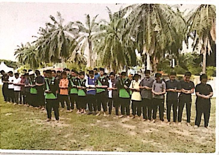 Murid menunaikan solat berjemaah di padang di bawah pokok kelapa